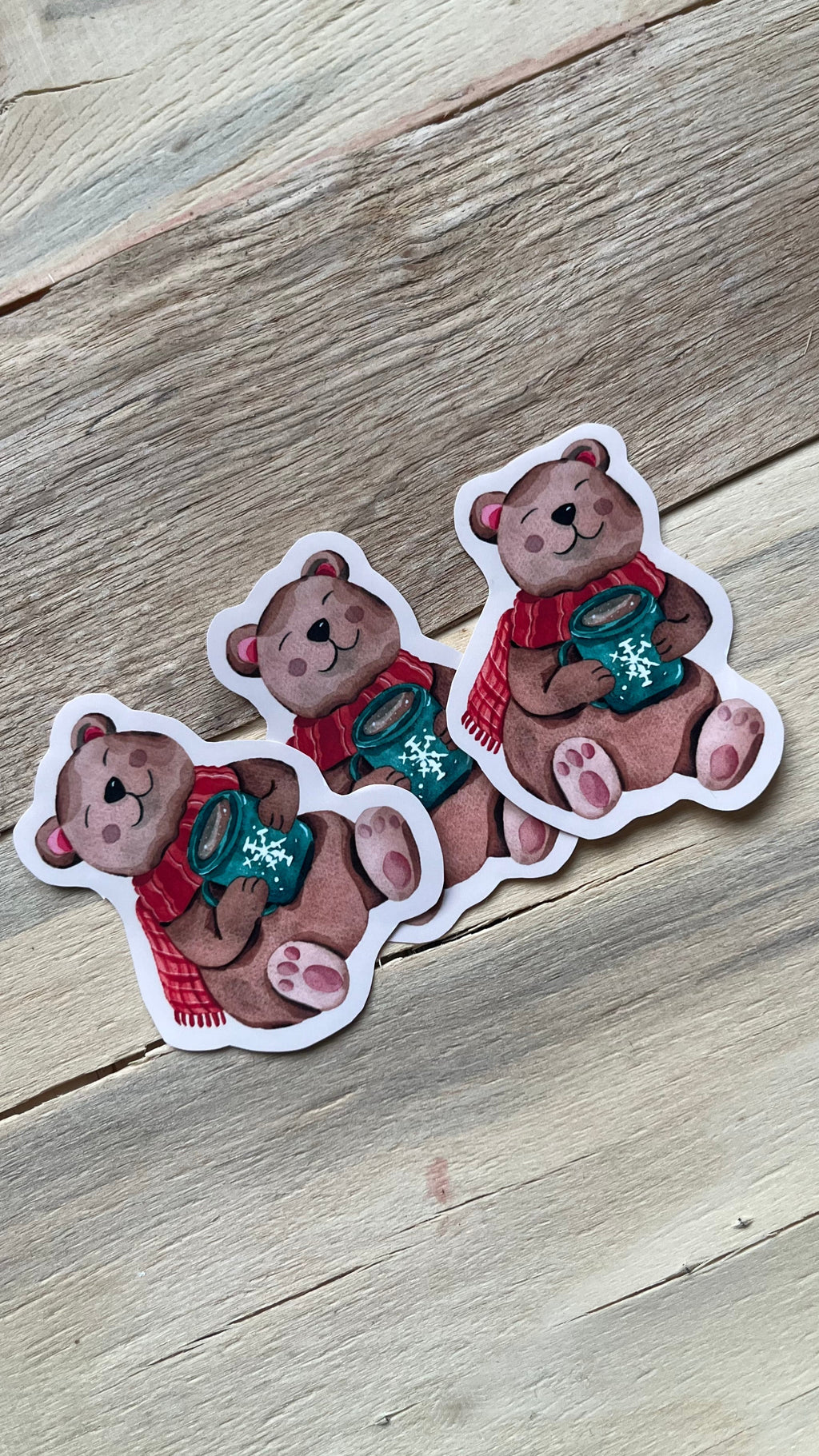 Trois stickers représentant un ours brun avec une écharpe rouge et une tasse verte décorée de flocons, sur un fond en bois clair.
