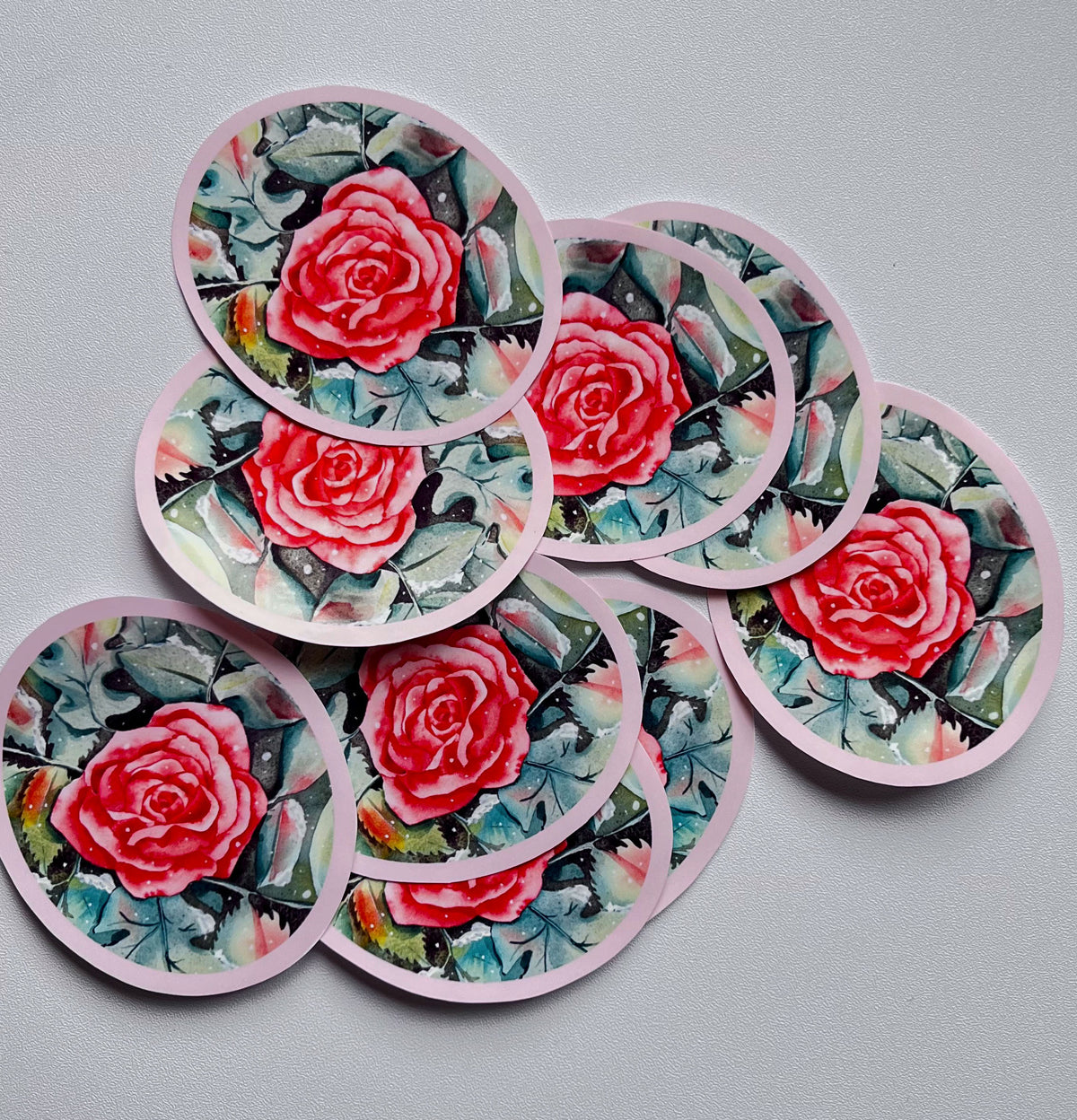 Stickers ronds Le Rosier d’hiver, ornés d’une rose rouge et rose sur un feuillage vert et bleu grisé, avec une bordure rose pâle, sur fond gris clair.