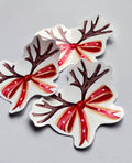 Trois stickers de Noël avec des bois de renne bruns, un nez rouge et un nœud rouge à étoiles blanches, photographiés sur un fond gris clair.