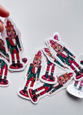 Stickers de Noël représentant un casse-noisette rouge et vert, avec une couronne dorée, une barbe blanche et des reflets étoilés, sur fond gris clair.