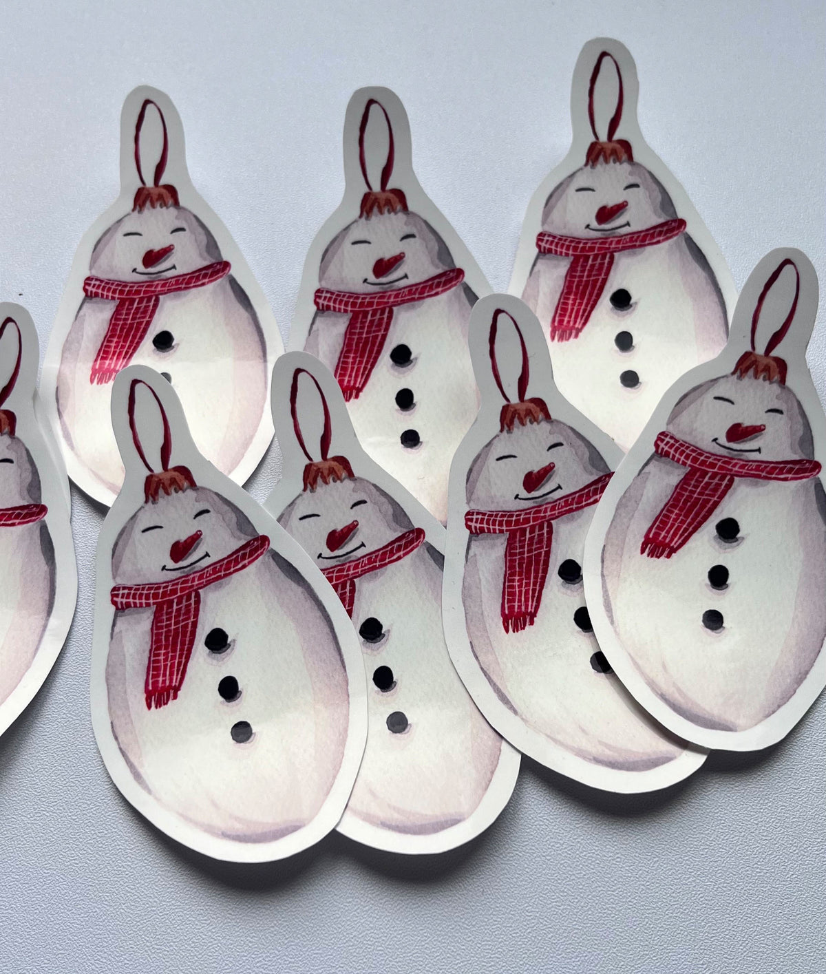 Stickers illustrés en forme de bonhomme de neige souriant avec une écharpe rouge et trois boutons noirs, disposés sur un fond gris clair.