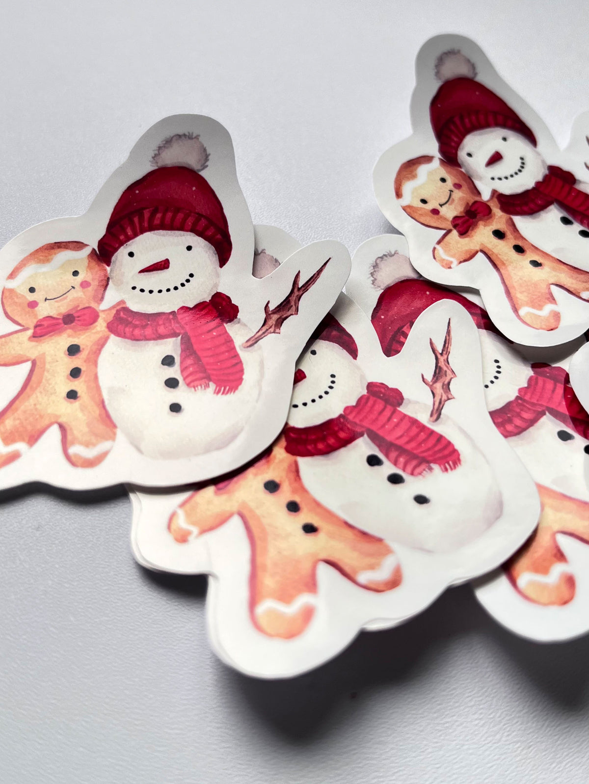 Stickers de Noël représentant un bonhomme de neige à bonnet rouge et écharpe rose aux côtés d’un bonhomme en pain d’épices, sur un fond gris clair.